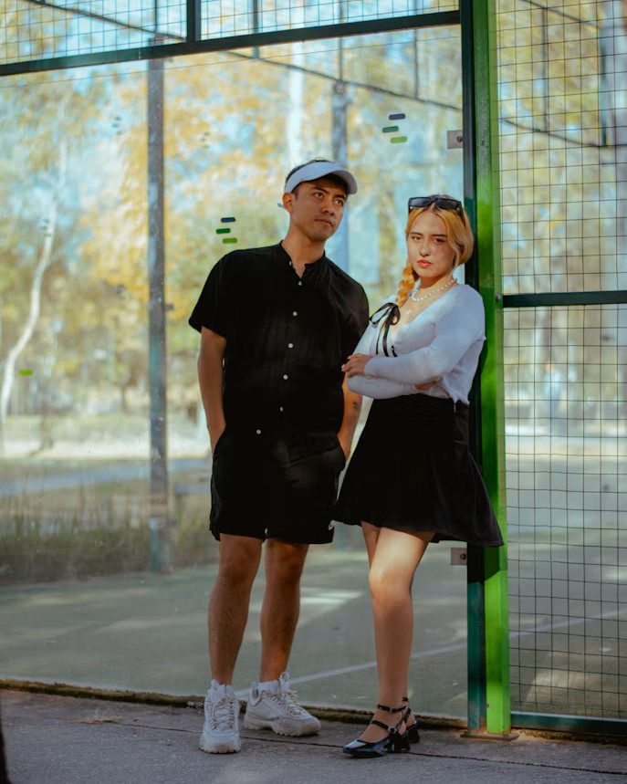 Een jonge man en vrouw in zwart-wit outfit poseren in de zon aan een padelterrein